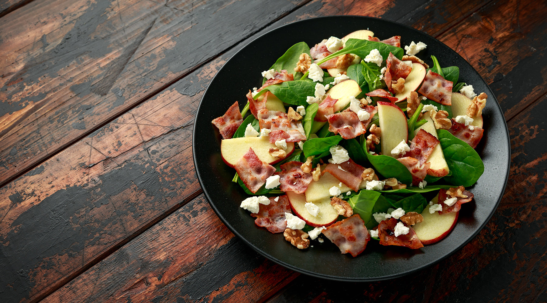 black bowl filled with green spring mix salad covered in apples. bacon, and goat cheese