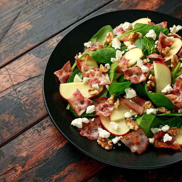 black bowl filled with green spring mix salad covered in apples. bacon, and goat cheese