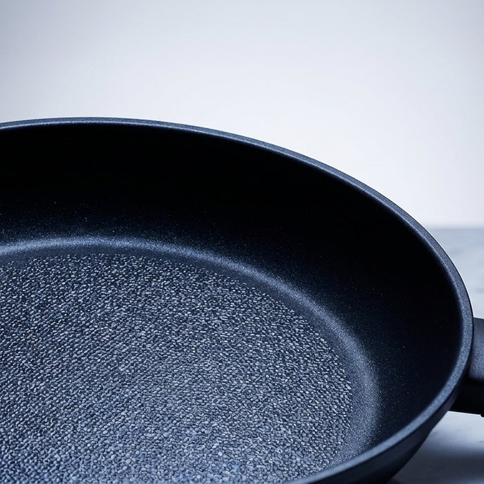 Close-up of a premium nonstick frying pan with a diamond-reinforced textured cooking surface, dramatically lit on a marble countertop against a soft grey background.